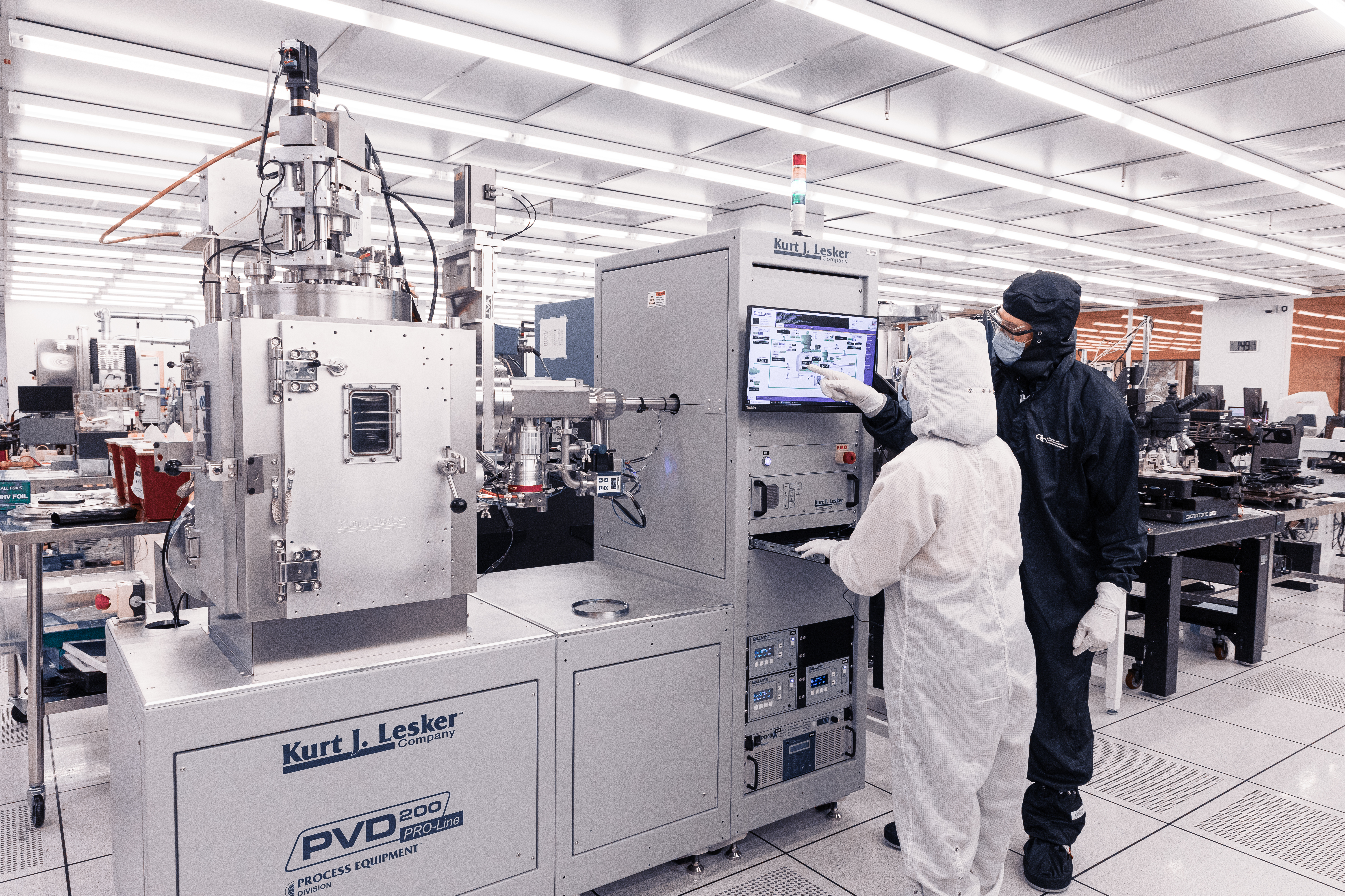 Researchers in cleanroom suits operate a Kurt J. Lesker deposition system inside a microelectronics fabrication facility.  One researcher points to the system’s control screen while another works at the equipment in a brightly lit cleanroom.