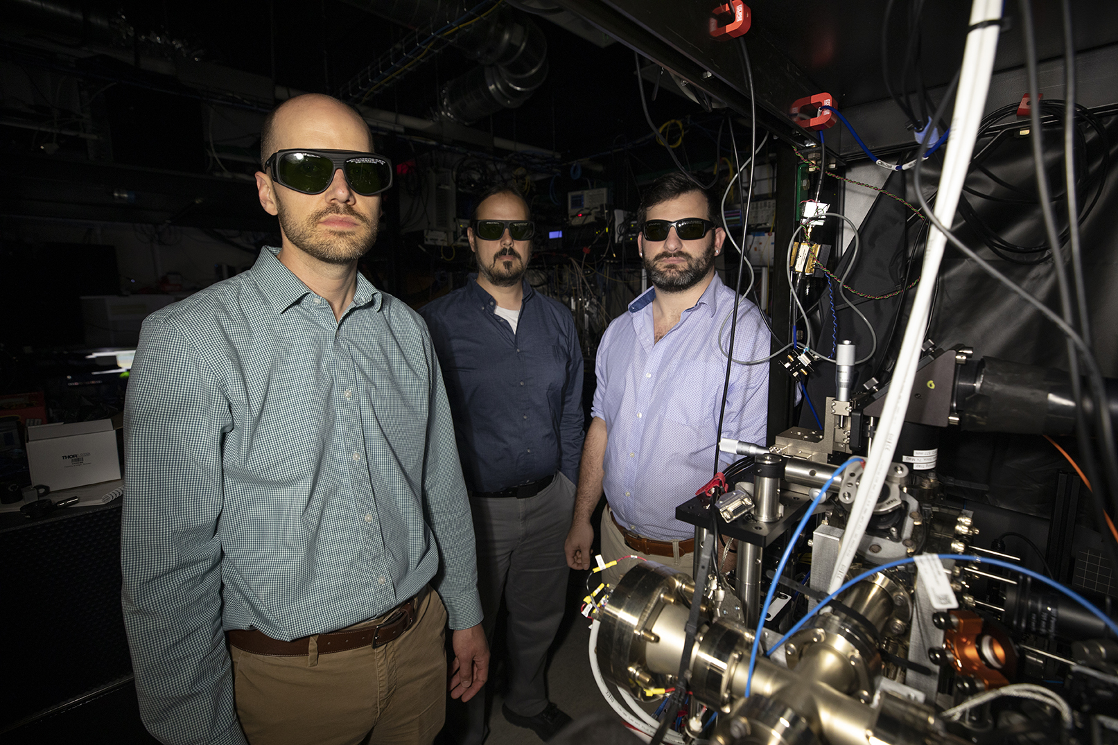 Researchers wearing laser safety glasses stand beside a complex quantum optics experiment featuring precision instrumentation, optical components, and interconnected cables in a laboratory setting.