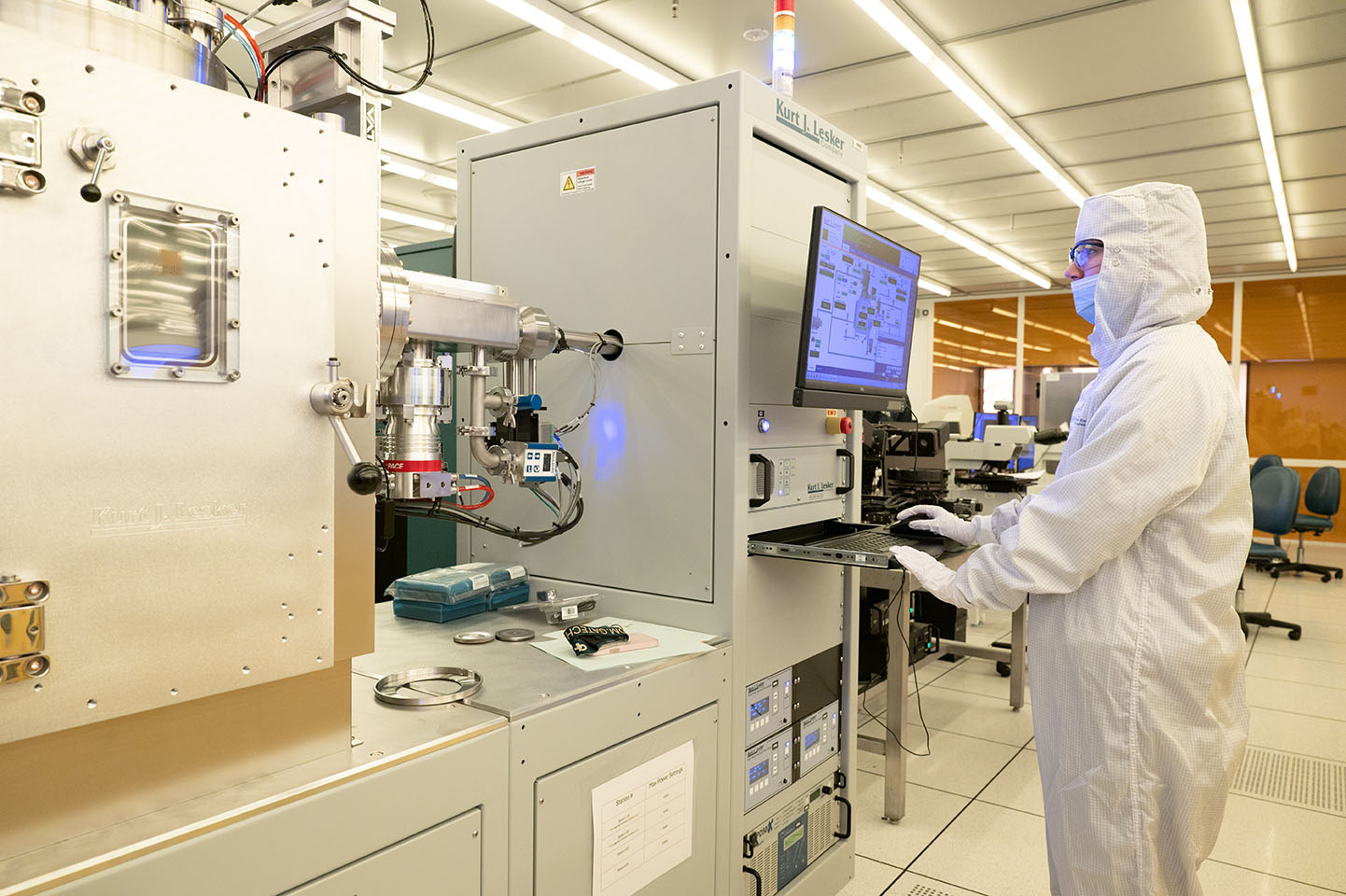 A cleanroom researcher operates a thin-film deposition system through a computer interface while monitoring advanced semiconductor manufacturing equipment.