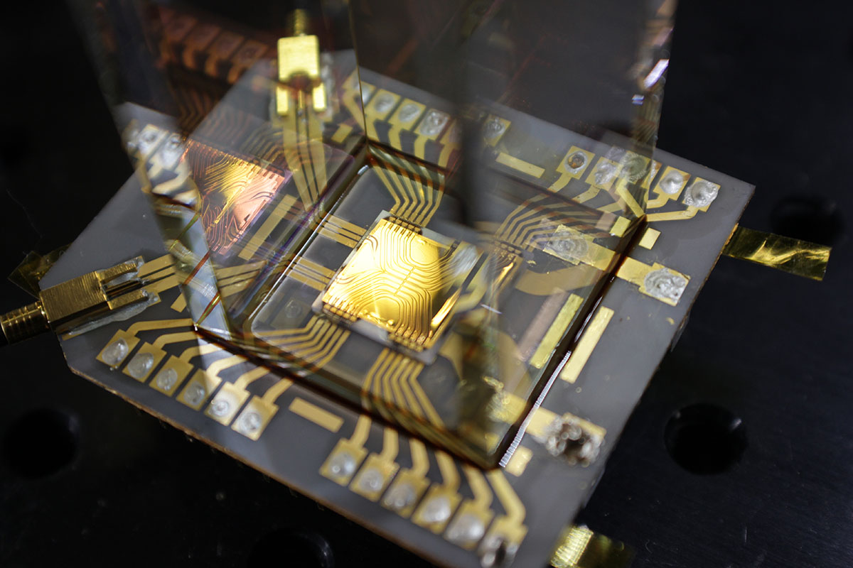 Close-up view of a microelectronic device mounted on a circuit board, with gold-colored conductive traces and precision probes connected for testing or measurement.