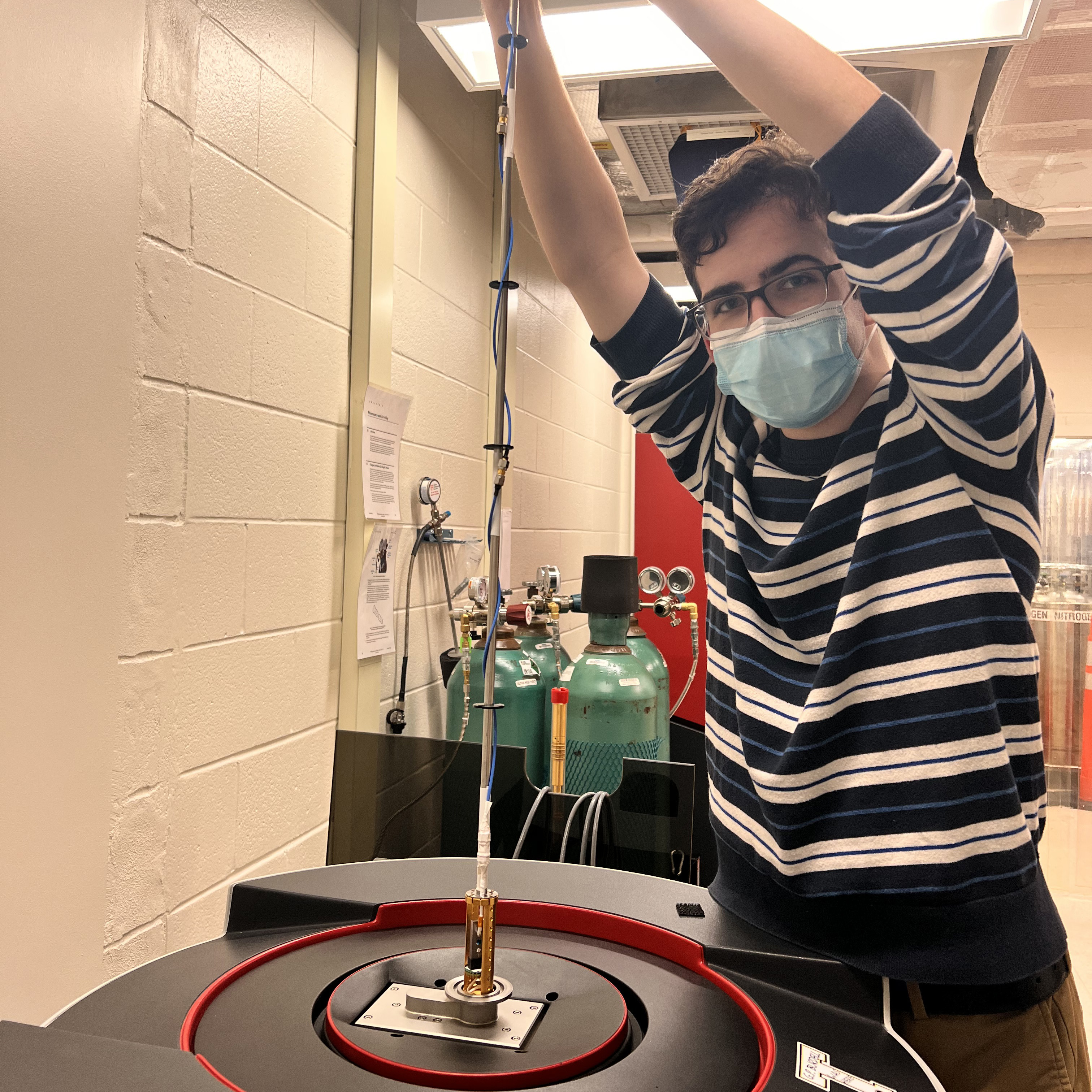 A student lowers a specialized probe into a cryogenic measurement system while preparing a materials science experiment in a laboratory.
