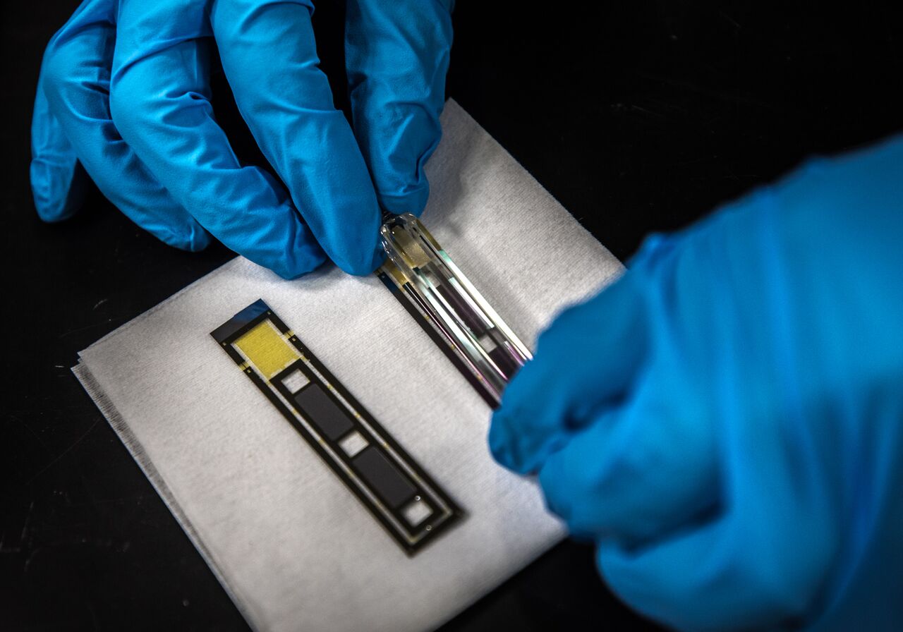 Gloved hands carefully handle a small electronic sensing device and transparent component during assembly or testing on a laboratory work surface.