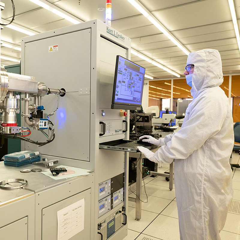 Researcher in cleanroom protective clothing operating semiconductor fabrication equipment in a laboratory.
