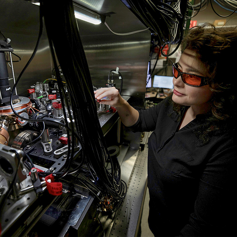 Researcher wearing protective glasses adjusts equipment in a laboratory filled with optical components, cables, and instrumentation.