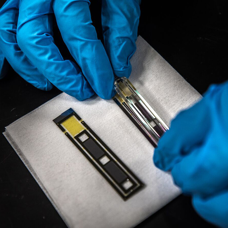Gloved hands handling a glass sensor device next to a microfabricated electronic chip on a laboratory work surface.