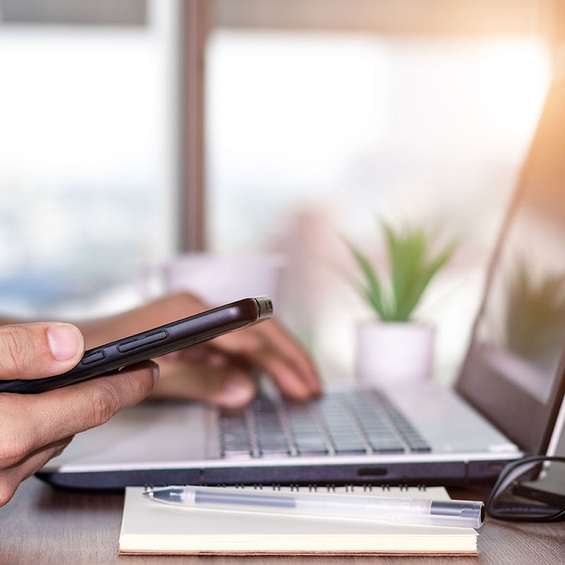 Hand holding a smartphone beside a laptop computer on a desk with a notebook and pen.