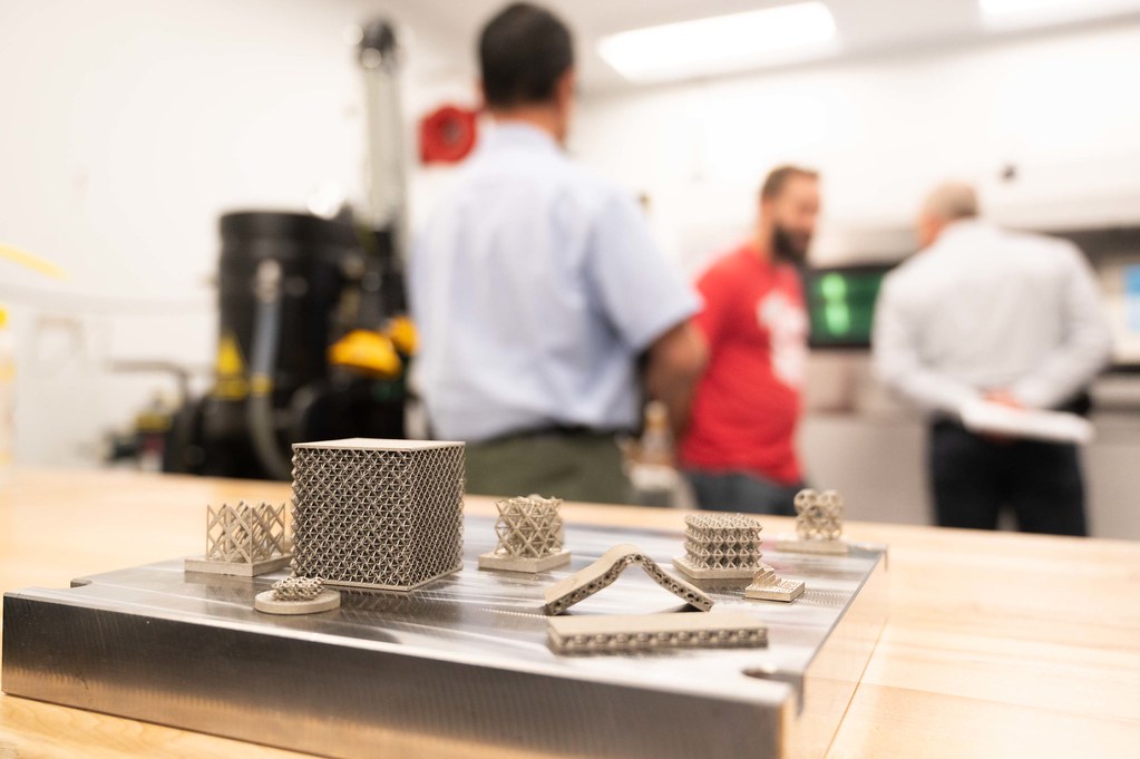 Close-up of metal 3D-printed lattice structures and geometric parts displayed on a metal plate in a workshop, with three people standing in the background near industrial equipment.