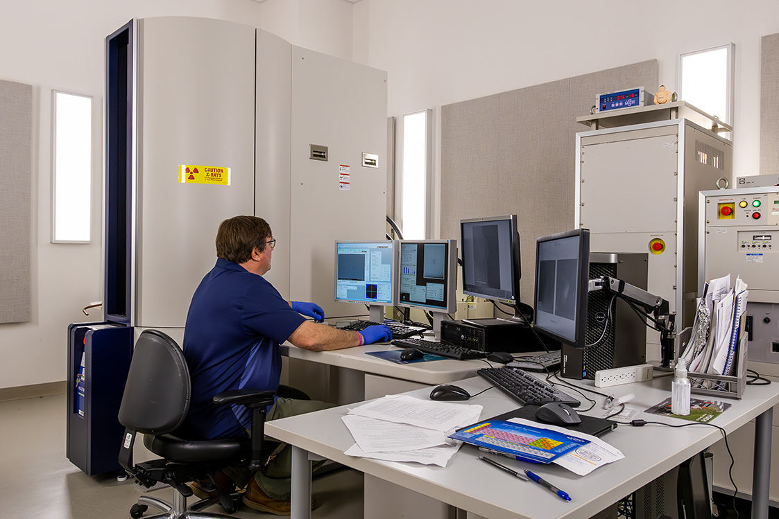 A researcher operates a materials characterization instrument while monitoring imaging and analysis software on multiple computer screens in a laboratory. 