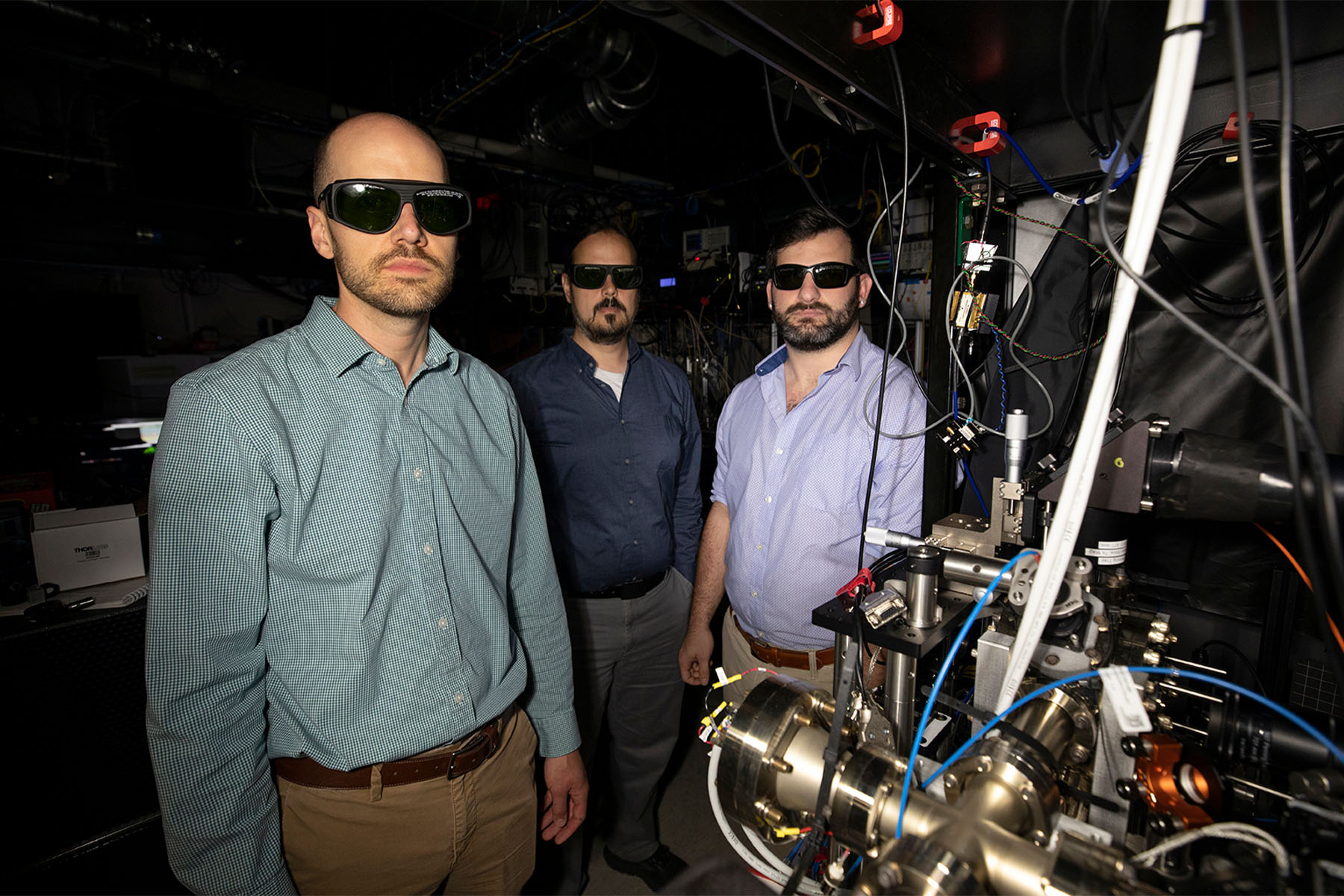 Researchers wearing laser safety glasses stand beside a complex quantum optics experiment featuring precision instrumentation, optical components, and interconnected cables in a laboratory setting.