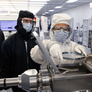 Two technicians in protective cleanroom clothing, masks, goggles, and gloves operate laboratory equipment, with one holding a circular metal component above an open chamber.