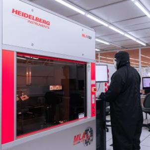A technician in protective cleanroom clothing operates a Heidelberg Instruments MLA150 maskless aligner in a brightly lit microfabrication laboratory.