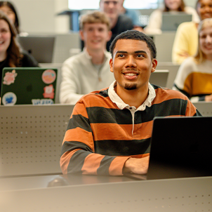 Student seated at a laptop in a classroom with other students in the background.