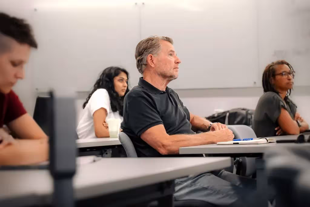 Adults seated at desks in a classroom, listening to a presentation or lecture during a training session.