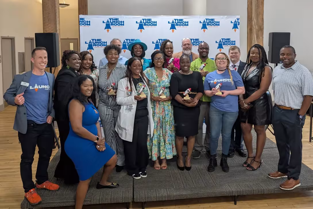 Group of award recipients and organizers pose with trophies in front of The Launch Room event backdrop.