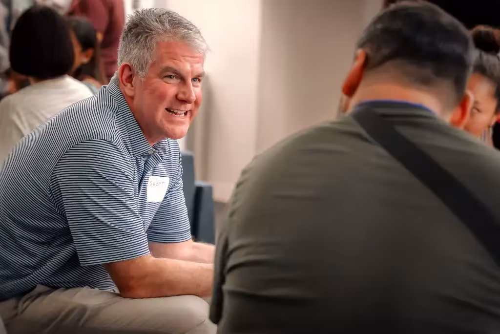 Participant in a striped shirt and name tag talks with others during a small group discussion at an indoor event.