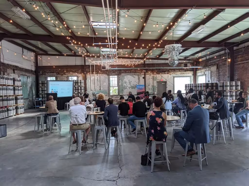 Attendees sit at tables facing a presentation screen in an industrial-style venue with string lights overhead.