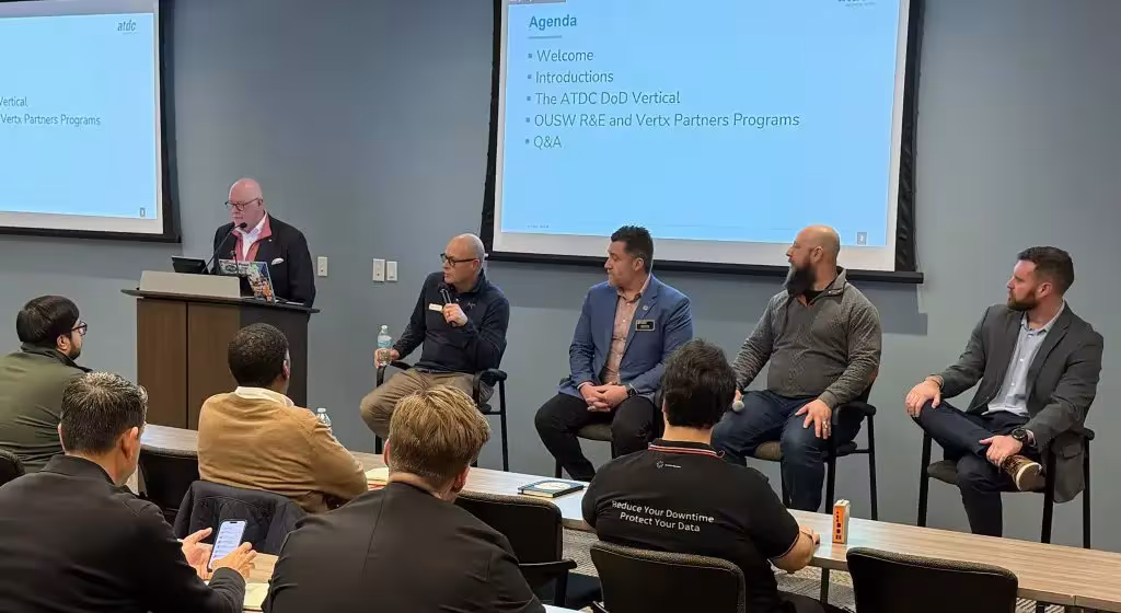 Panel discussion with five speakers addressing attendees in a conference room beneath agenda slides.