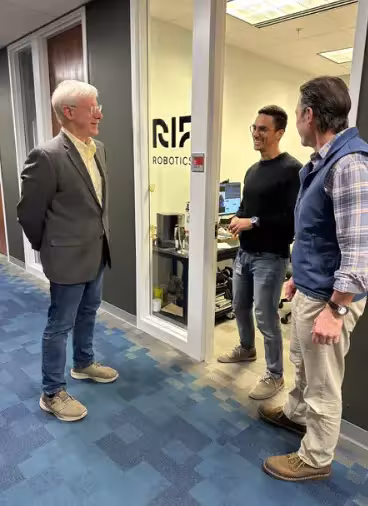 Three attendees stand outside a robotics startup office, talking in a hallway at an innovation workspace.